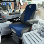 Interior view of a modern tractor cab with blue and gray seat, control panel, joystick, and protective plastic covers; outside show tractors and a brick building.