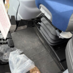Footwell area inside a bus or coach: blue seat, gray plastic trim, exposed pedals and seat adjustment lever, with loose plastic bags on the floor nearby.