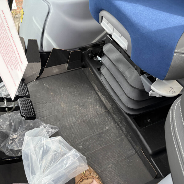 Footwell area inside a bus or coach: blue seat, gray plastic trim, exposed pedals and seat adjustment lever, with loose plastic bags on the floor nearby.
