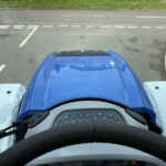 View from the driver’s seat in a blue vehicle, showing the steering wheel and dashboard with road and dashed lane markings ahead.