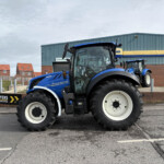 Two large blue New Holland tractors parked in a lot near a blue industrial building and brick wall at a commercial site.