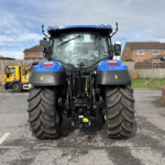 Front view of a large blue tractor parked on a suburban street, with a yellow truck and brick houses in the background.