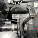 Close-up of a dusty vehicle engine area showing belts, hoses, clamps, and a metal identification plate with small text.