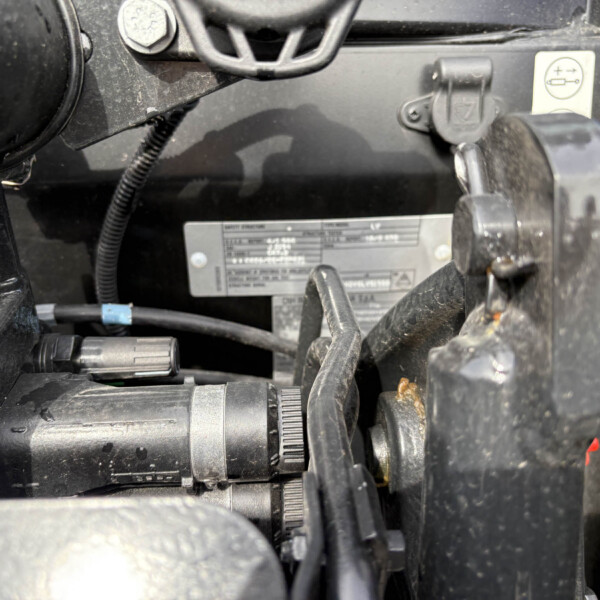 Close-up of a dusty vehicle engine area showing belts, hoses, clamps, and a metal identification plate with small text.