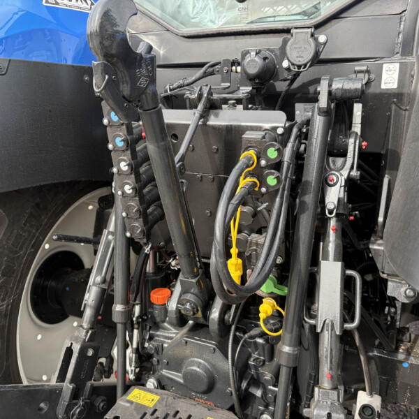 Close-up of a tractor's front-end hydraulic system with hoses, valves, and linkages beside a blue body panel