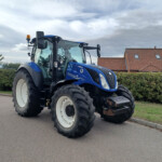 Blue New Holland tractor parked on a street with hedges and a red-tiled house behind it under a cloudy sky