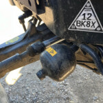 Close-up of a dusty trailer hitch ball and mounting bracket on a vehicle frame, with a triangular ID sticker reading BK8X nearby.