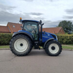 Blue tractor parked on a street with red brick houses in the background and cloudy sky above.