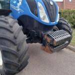 Front view of a blue New Holland tractor with large rear tires and rusted metal steps, parked on a driveway with hedges in the background.