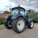Front view of a large blue tractor with oversized rear tires parked on a road, orange warning lights on top, and cloudy sky overhead.
