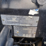 Close-up of a metal data plate on a dusty black surface, showing safety/structure information and VIN numbers partially obscured by scratches.