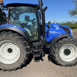 Blue New Holland tractor shown from the side, with large front tires and an orange beacon on top near a barn-like structure