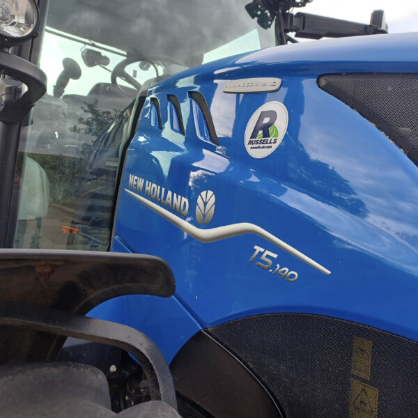 Close-up of a blue New Holland tractor side with dealership sticker, showing the 'RUSSELLS' badge and 'NEW HOLLAND' branding on the cab area.