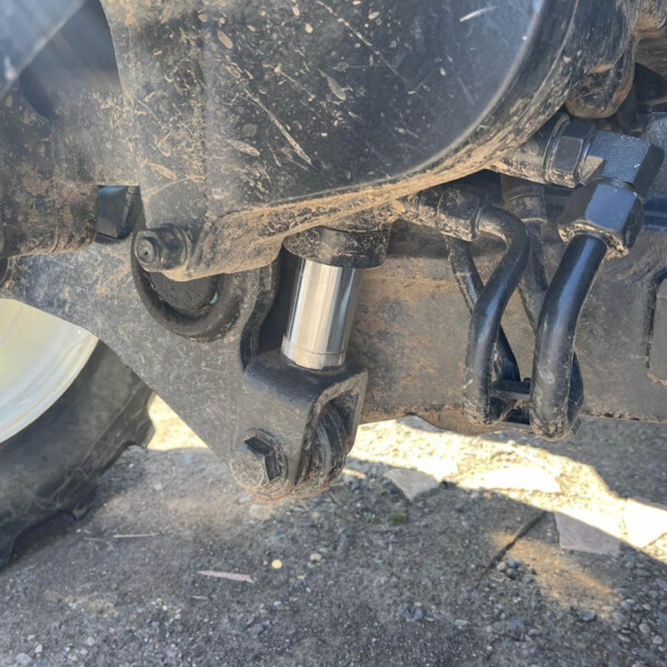 Close-up of a dirt-covered motorcycle/trailer chassis underside showing bolts, a hydraulic line, and a cylindrical shock absorber near the wheel.” Load-bearing components and muddy surface visible.