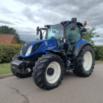Blue New Holland tractor parked on a street with hedges and a red-tiled roof house in the background under a cloudy sky.