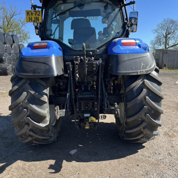 Rear view of a blue agricultural tractor with oversized tires, orange beacon lights, and a visible hitch at the back on a dirt surface, plate KV72 HJN on the left side.
