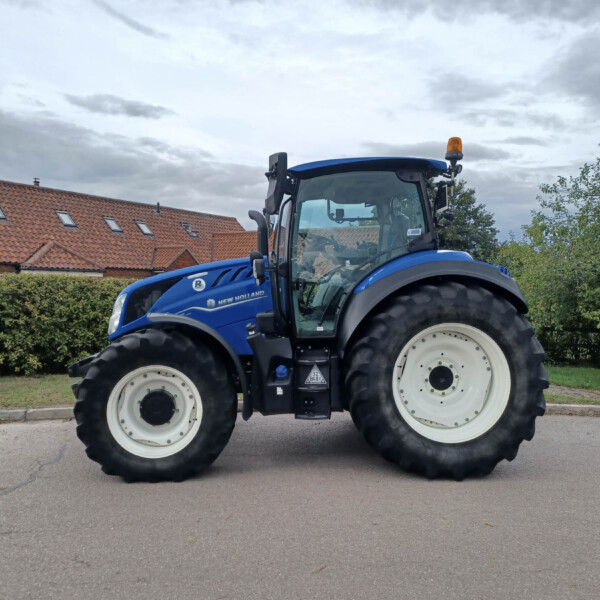 Blue New Holland tractor parked on a residential street with a red-tiled house and hedges in the background.