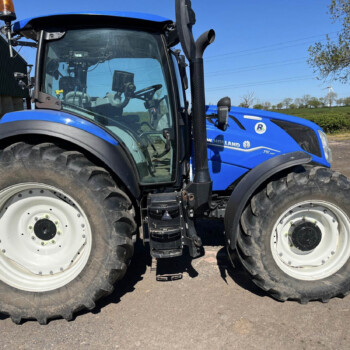 Blue New Holland tractor shown from the side, with large front tires and an orange beacon on top near a barn-like structure