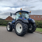 Blue agricultural tractor parked on a street with red-tiled buildings and trimmed hedges in the background.