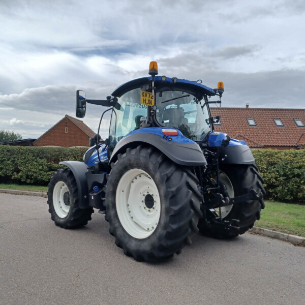 Blue agricultural tractor parked on a street with red-tiled buildings and trimmed hedges in the background.