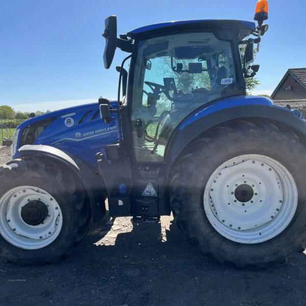 Close-up side view of a blue New Holland tractor with large front tires on a dirt surface, sunny day, rural yard in background.