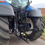 Front-left view of a blue tractor showing hydraulic levers and exposed machinery near a large rear tire.