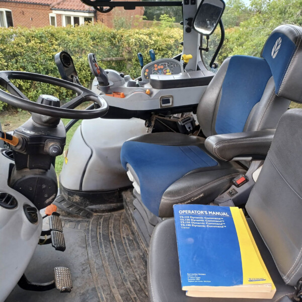 View of a tractor cab interior showing a large steering wheel, foot pedals, and control panel with colored switches; blue and gray seat in foreground with an operator's manual on the seat edge.