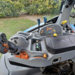 Inside the cab of a blue tractor showing the control panel with colored buttons, levers, and a side mirror, with greenery outside the window.