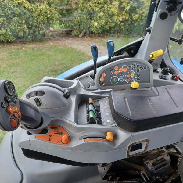 Inside the cab of a blue tractor showing the control panel with colored buttons, levers, and a side mirror, with greenery outside the window.
