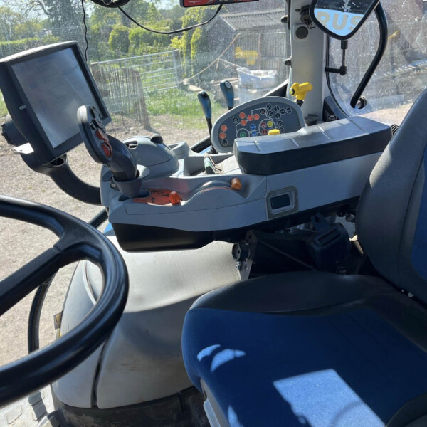 Cockpit of a farm tractor with a touchscreen display and a cluster of colored control buttons, blue seat, and dusty windshield.