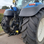 Close-up of a blue agricultural tractor's front end, showing large rugged tires and exposed hydraulic components.