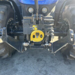 Close-up of a blue tractor front with hitch, hydraulic components, and a bright yellow PTO cover between two large tires.