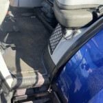 Driver's cockpit of a blue vehicle showing the black leather seat, pedals (gas and brake) and a New Holland floor mat.