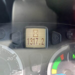 Car dashboard viewed from driver’s seat; center display shows an hourglass icon and numbers; red brake warning light on; gear indicator shows N.