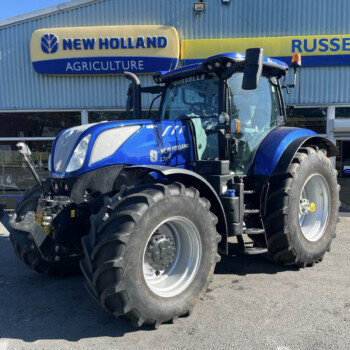 Blue New Holland tractor outside an agriculture shop, dealership signs reading 'New Holland Agriculture' in the background.