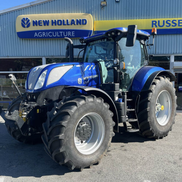 Blue New Holland tractor outside an agriculture shop, dealership signs reading 'New Holland Agriculture' in the background.