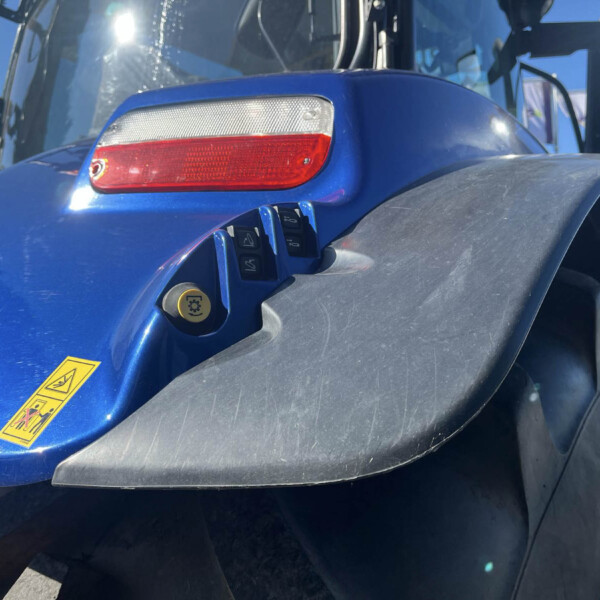 Close-up of a blue tractor hood showing a red rear light, control switches, and yellow warning stickers on a dusty surface.