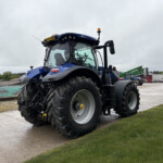 Blue agricultural tractor pictured from the front-left, with oversized tires in a yard of farming equipment on a cloudy day.