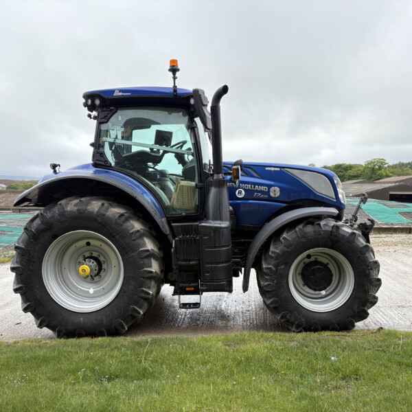 Blue New Holland tractor with an orange beacon on top sits on a gravel lot under a cloudy sky, large tires visible in foreground.