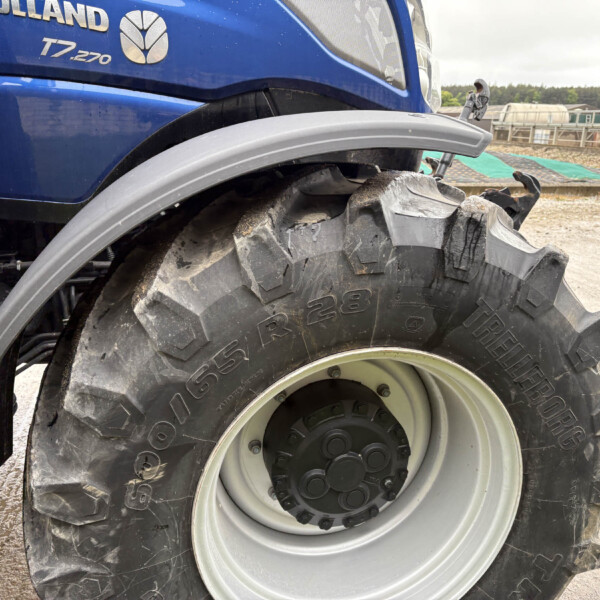 Close-up of a large tractor tire and blue body, showing thick tread, wheel rim, and muddied ground near a farm setting.