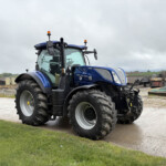 Blue New Holland tractor parked in a farmyard with green grass in the foreground and cloudy sky above.