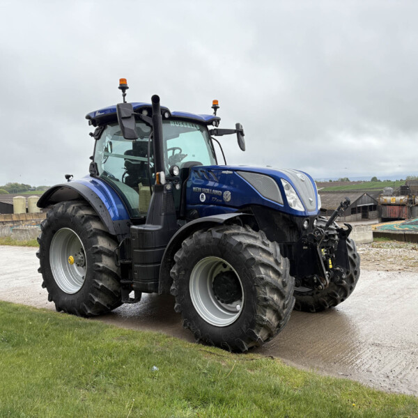 Blue New Holland tractor parked in a farmyard with green grass in the foreground and cloudy sky above.