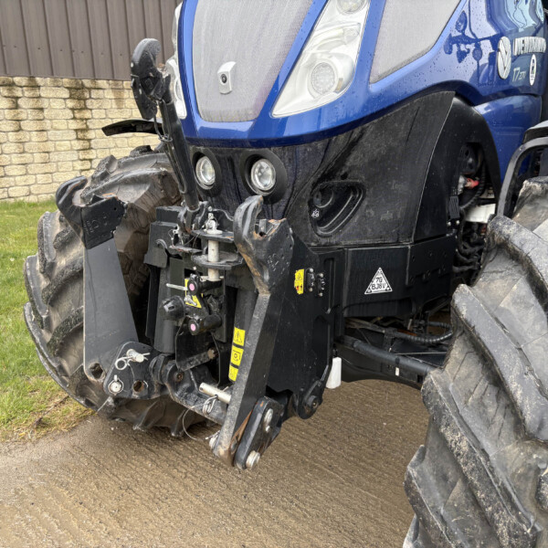 Blue tractor with muddy tires and damaged front end, parked on a grassy area beside a brick wall.