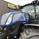 Blue New Holland tractor front view with mud on tires parked beside a brown building with a New Holland Agriculture sign in the background