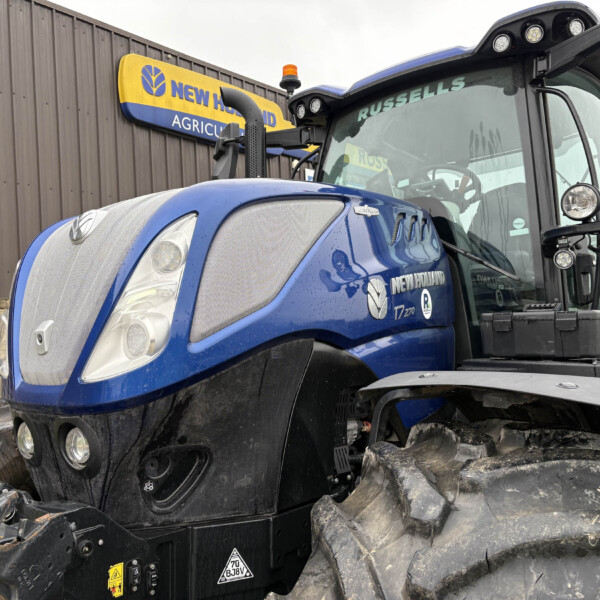 Blue New Holland tractor front view with mud on tires parked beside a brown building with a New Holland Agriculture sign in the background