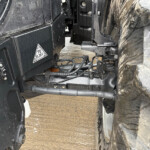 Close-up of heavy machinery linkage and hydraulic hoses between a black machine and a muddy tire, with warning labels visible.