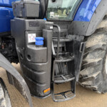 Close-up of a blue tractor cab with mud-splattered steps, large front tire, and a blue fuel cap visible near the steps.