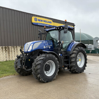 Blue New Holland agricultural tractor with large tires parked beside a dark industrial building and a yellow-blue New Holland Agriculture sign above it