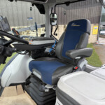 Interior view of a tractor cab with a blue and gray driver seat, steering wheel, and a tablet on the left; view shows an industrial yard outside the windows.