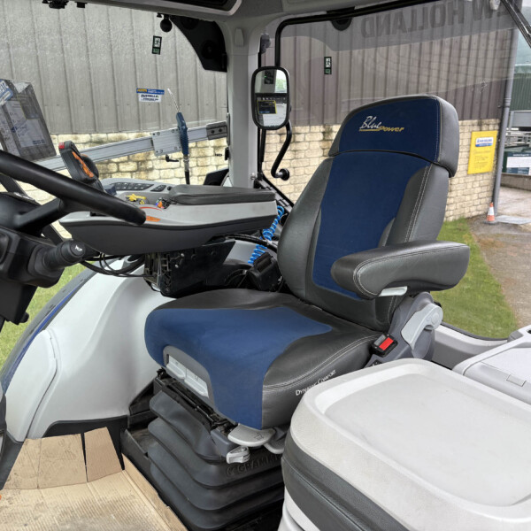 Interior view of a tractor cab with a blue and gray driver seat, steering wheel, and a tablet on the left; view shows an industrial yard outside the windows.